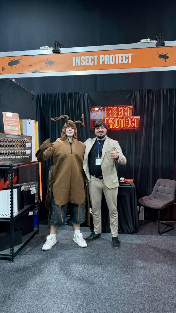 Two men at an exhibition booth with an 'INSECT PROTECT' sign; one in a brown animal costume and the other in a beige suit with a badge, giving a thumbs up.