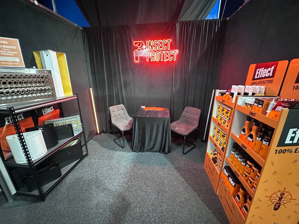 Booth corner with black drapes, a red neon 'INSECT PROTECT' sign, and two gray chairs around a small table.