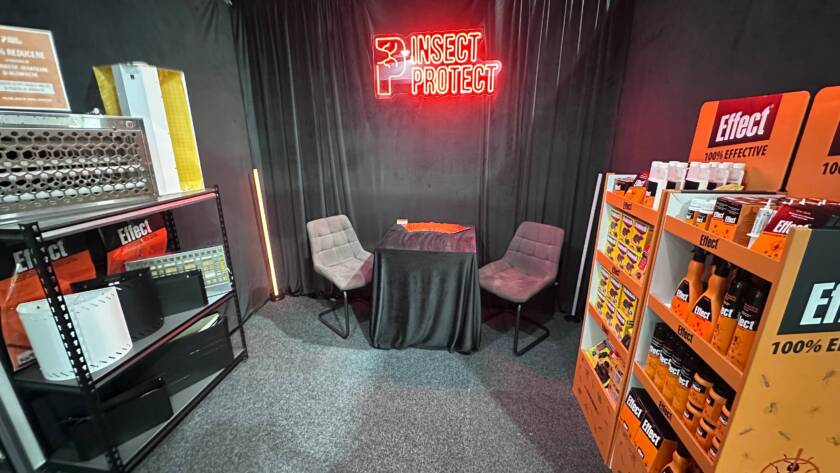 Booth corner with black drapes, a red neon 'INSECT PROTECT' sign, and two gray chairs around a small table.